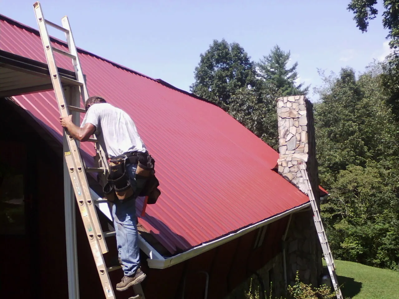 Roofer on a ladder beside a red metal roof and stone chimney — Soffit & Fascia Repair by Roofing Brothers in Yatesboro